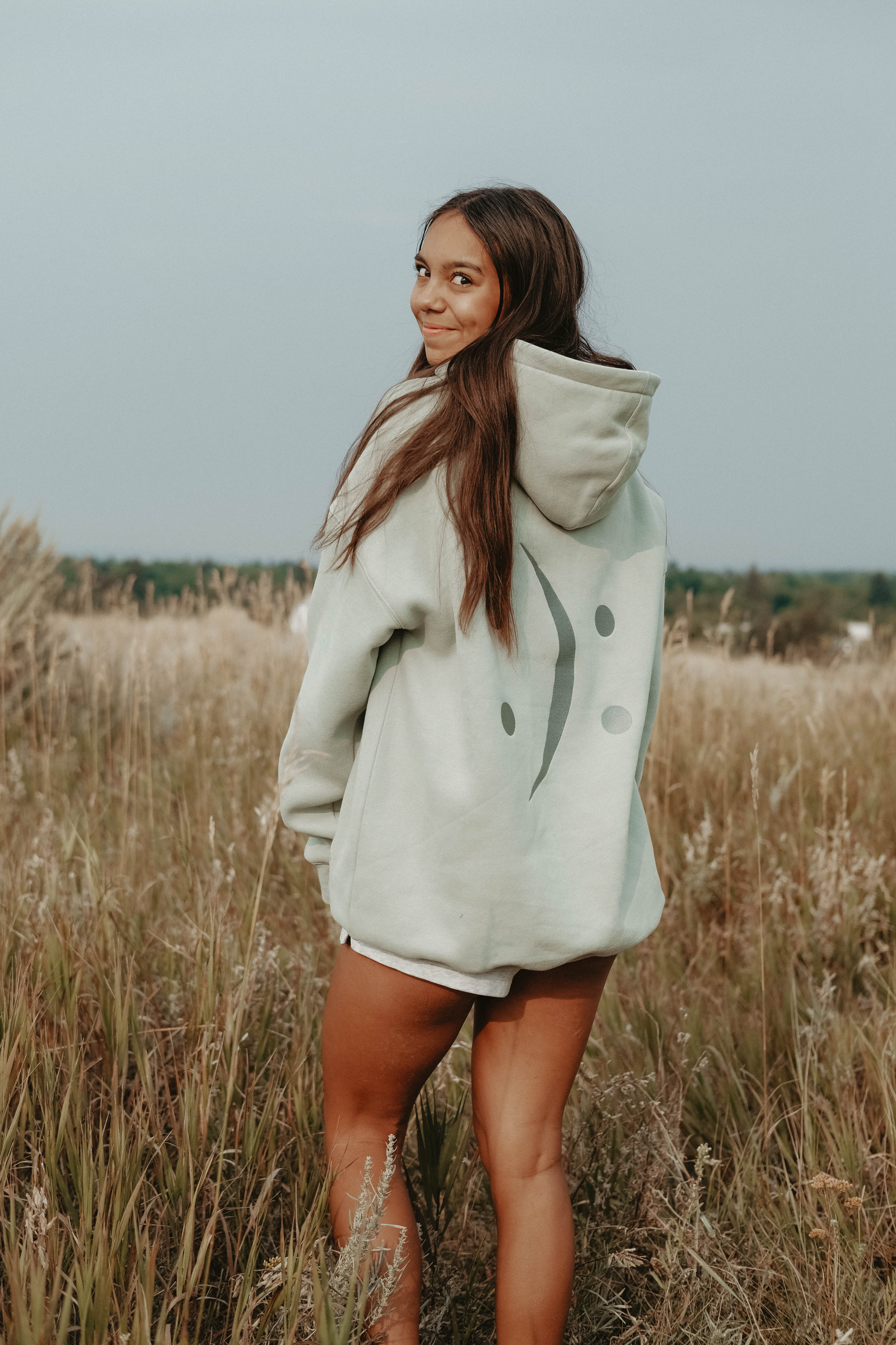 A woman standing in a field of dry grass wearing a light sage green oversized hoodie with a dark gray smiley face graphic on the front.