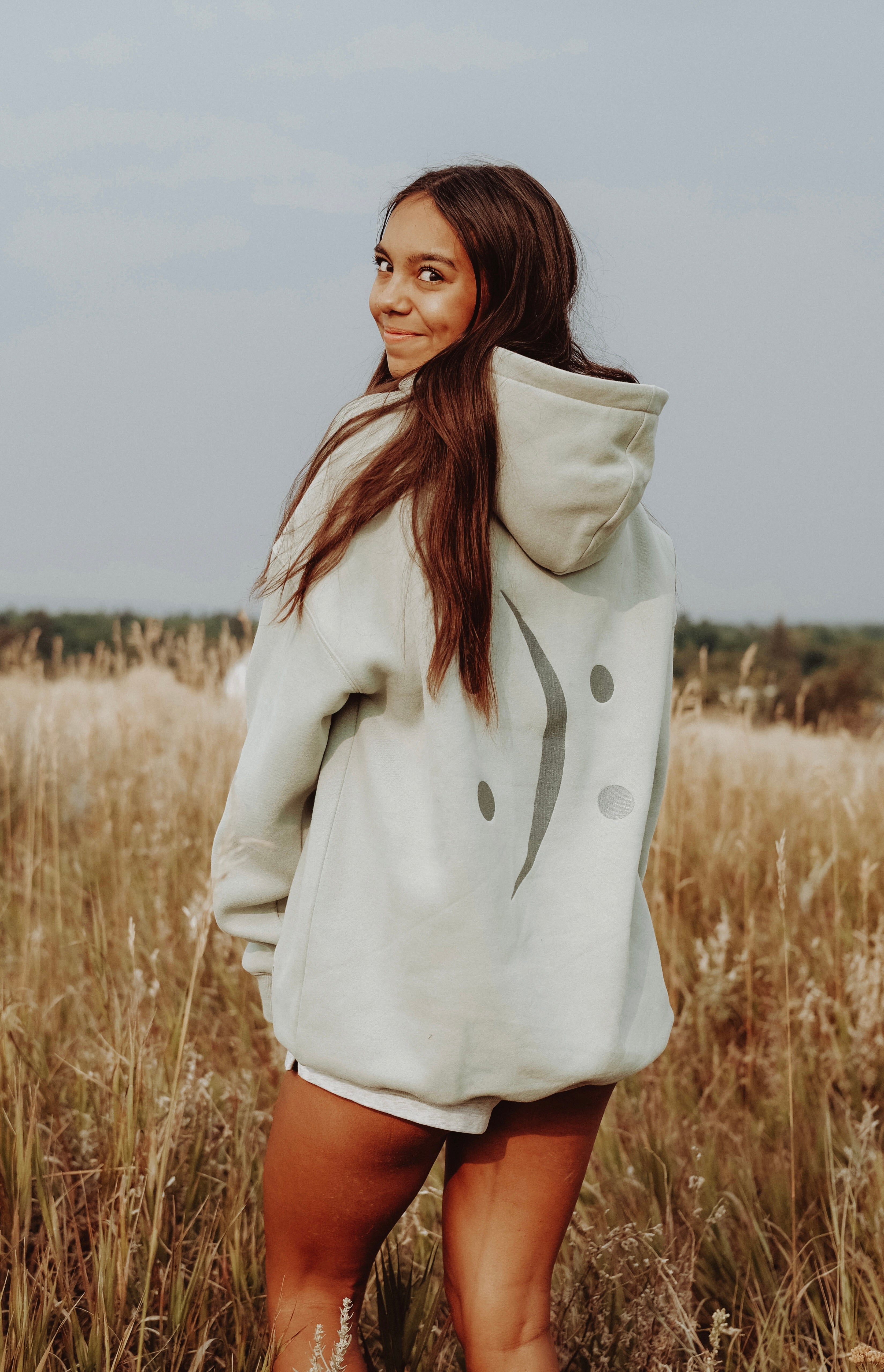 A woman standing in a field of dry grass wearing a light sage green oversized hoodie with a dark gray smiley face graphic on the front.
#color_matcha