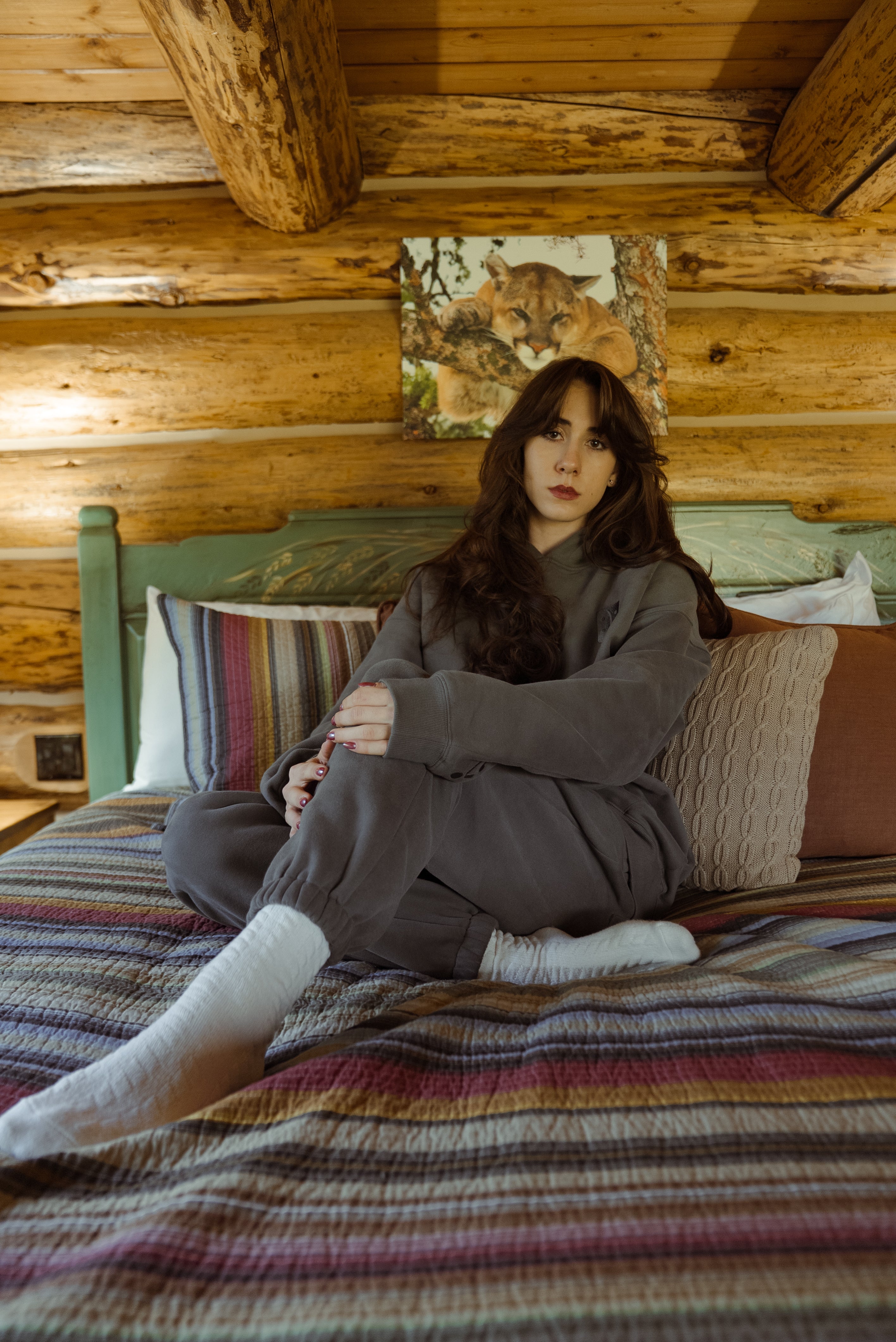 A woman sits cross-legged on a bed in a rustic log cabin bedroom, wearing a dark gray lounge set and white socks. The bed has a striped multicolor blanket, and the background features wooden walls and a green headboard.