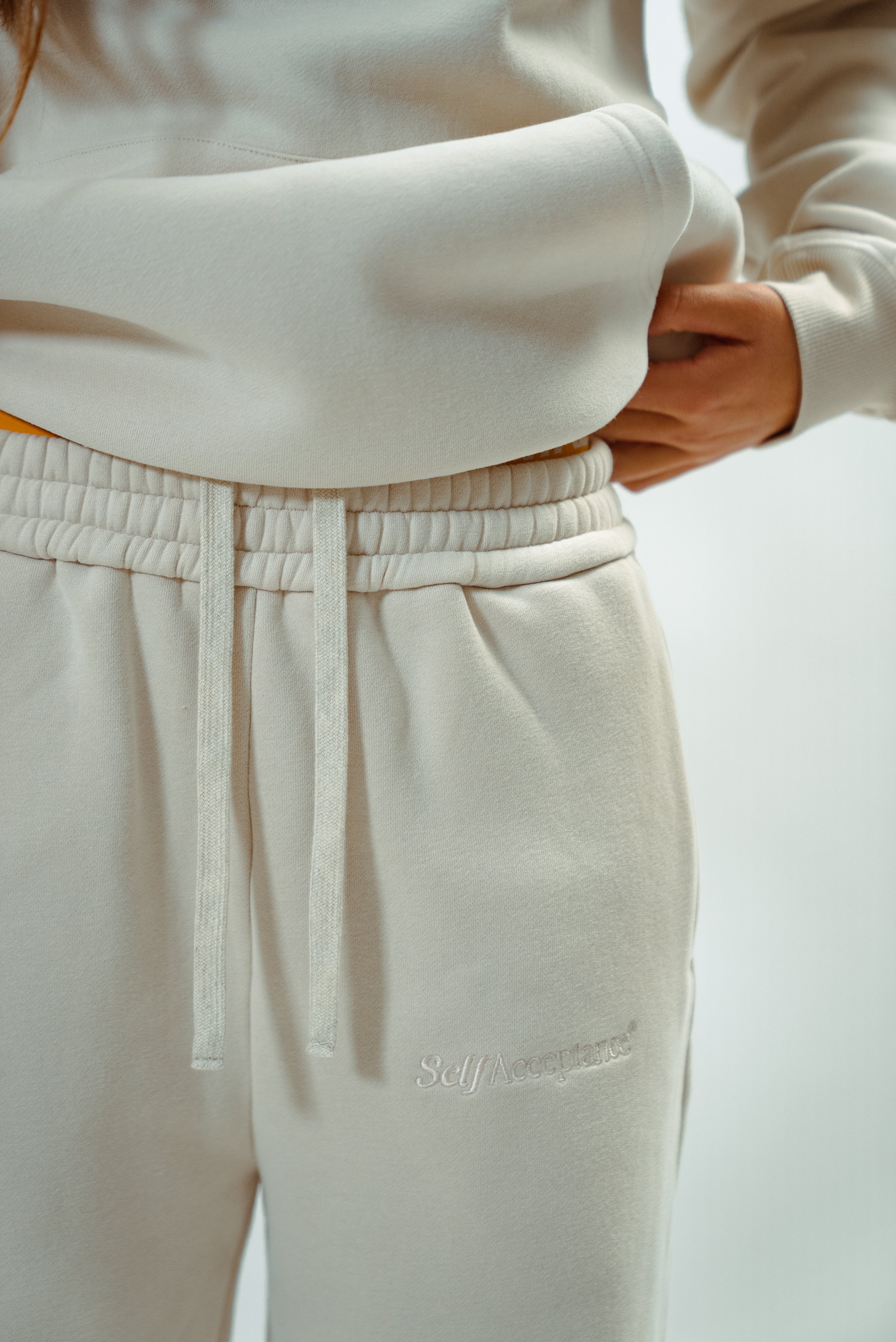 Close-up of cream Self Acceptance™ sweatpants with a drawstring waist and tonal embroidered logo, paired with a matching hoodie.