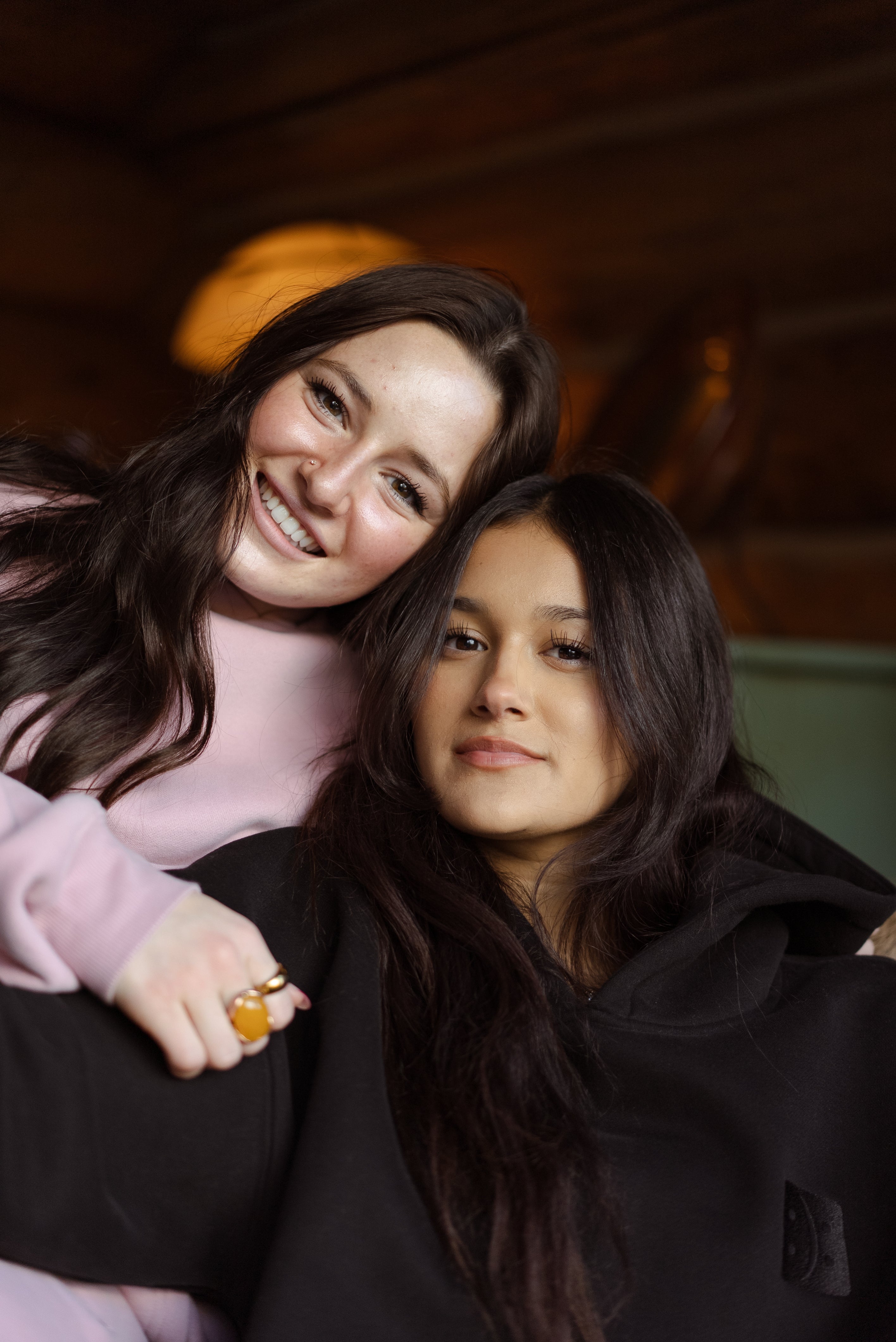 Two women wearing pink and black Self Acceptance™ hoodies pose together and smile indoors.