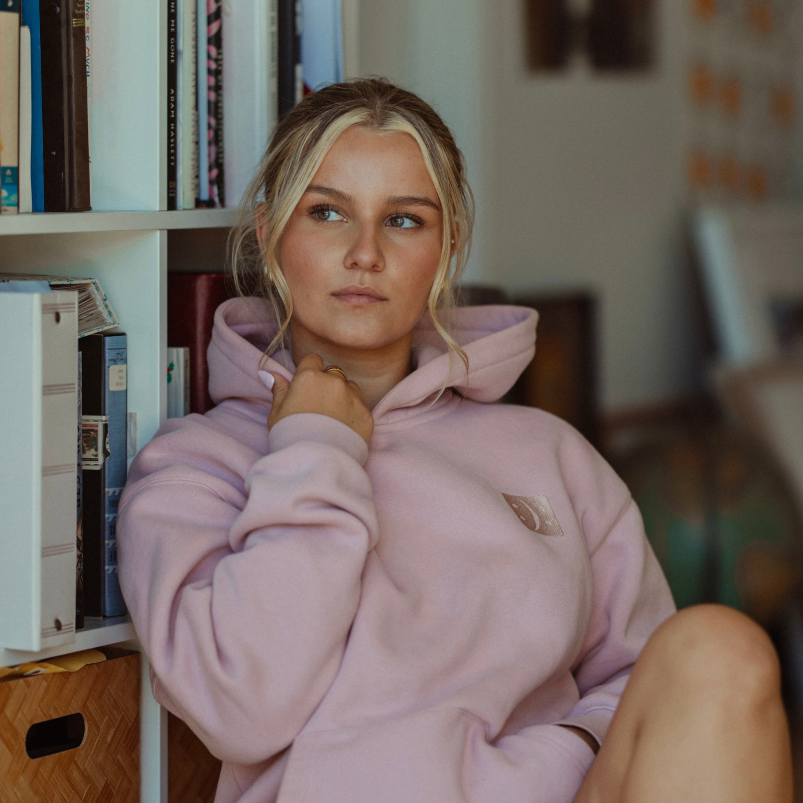 A woman with blonde hair sitting in front of a bookshelf, wearing a light pink oversized hoodie sweatshirt with a small embroidered logo on the chest.