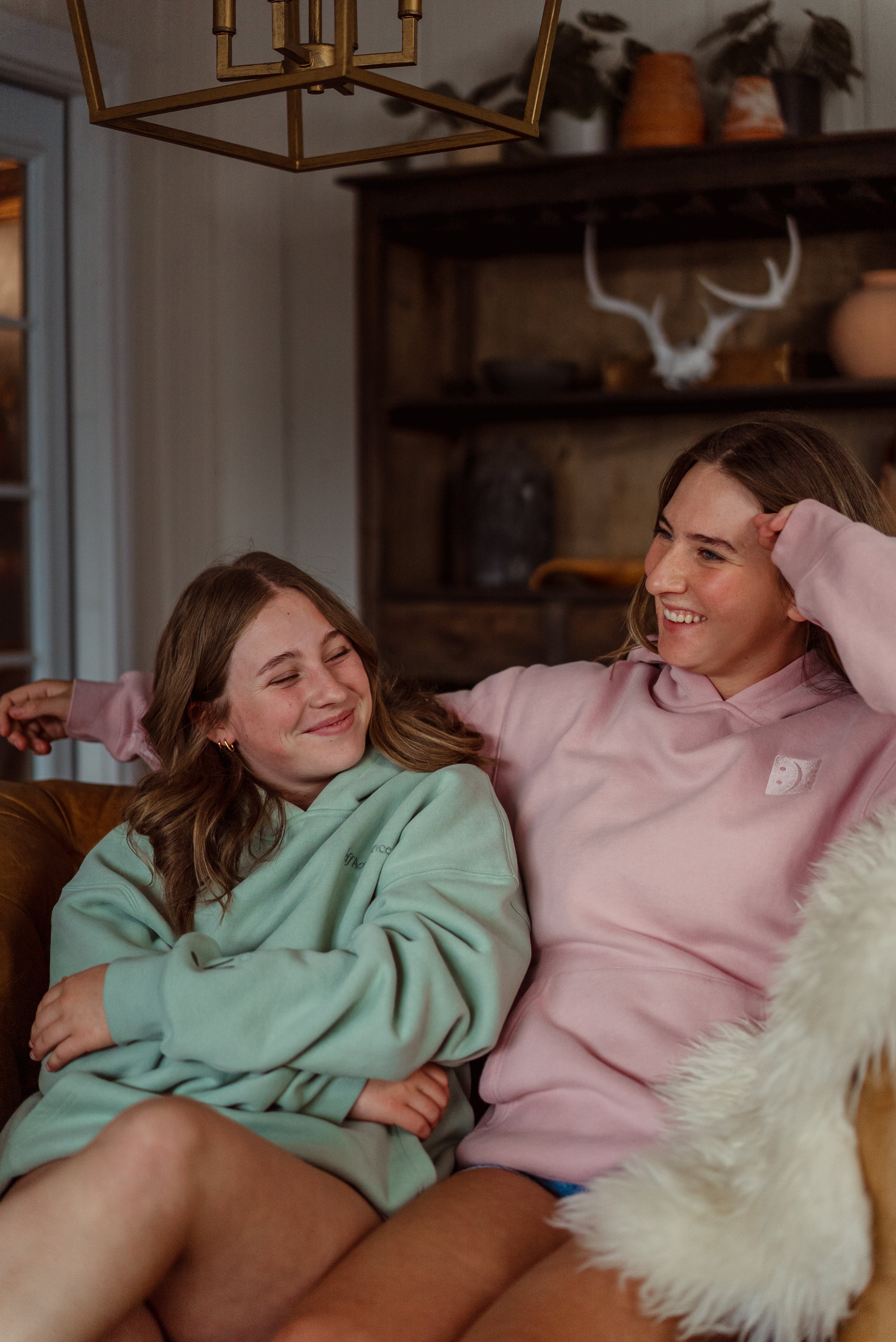 Two women sitting closely together on a couch in a cozy living room setting. The woman on the left wears a light sage green oversized hoodie, and the woman on the right wears a dusty pink oversized hoodie with a small white square logo on the left chest.