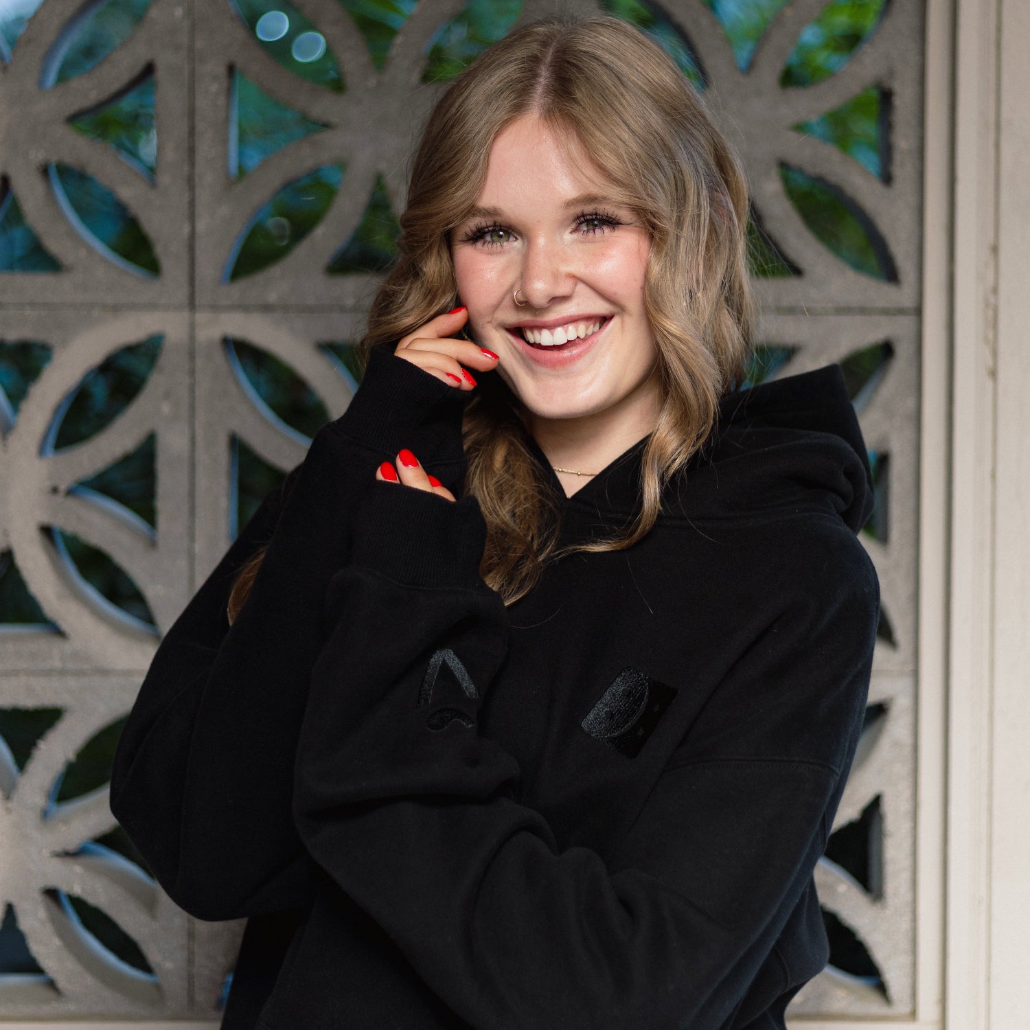 A smiling young woman with blonde hair wearing a black hoodie, posing in front of a decorative stone screen.