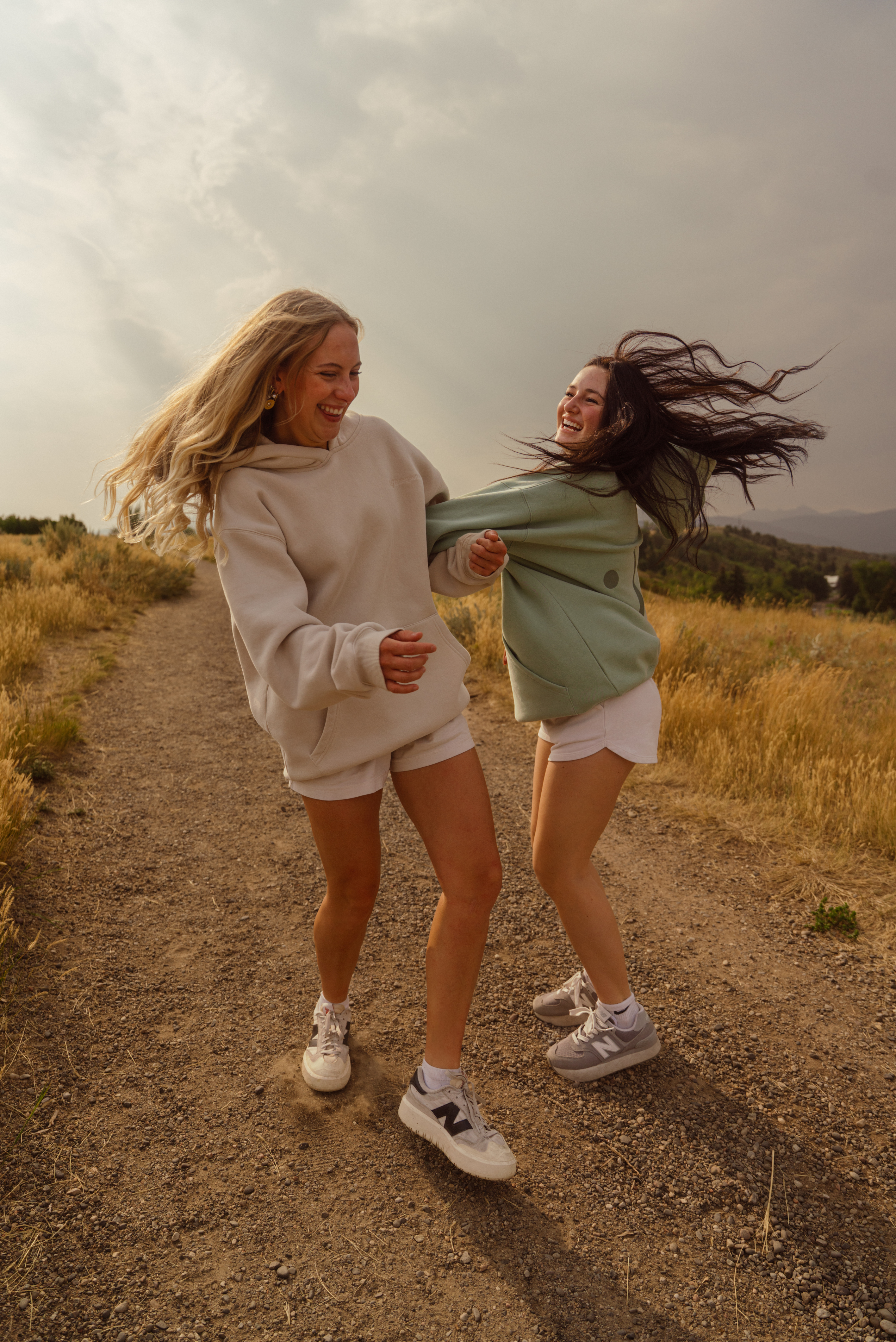 Two women laughing and playfully spinning on a dirt path outdoors, wearing matching oversized sweatshirts in cream and sage green with white shorts and sneakers.