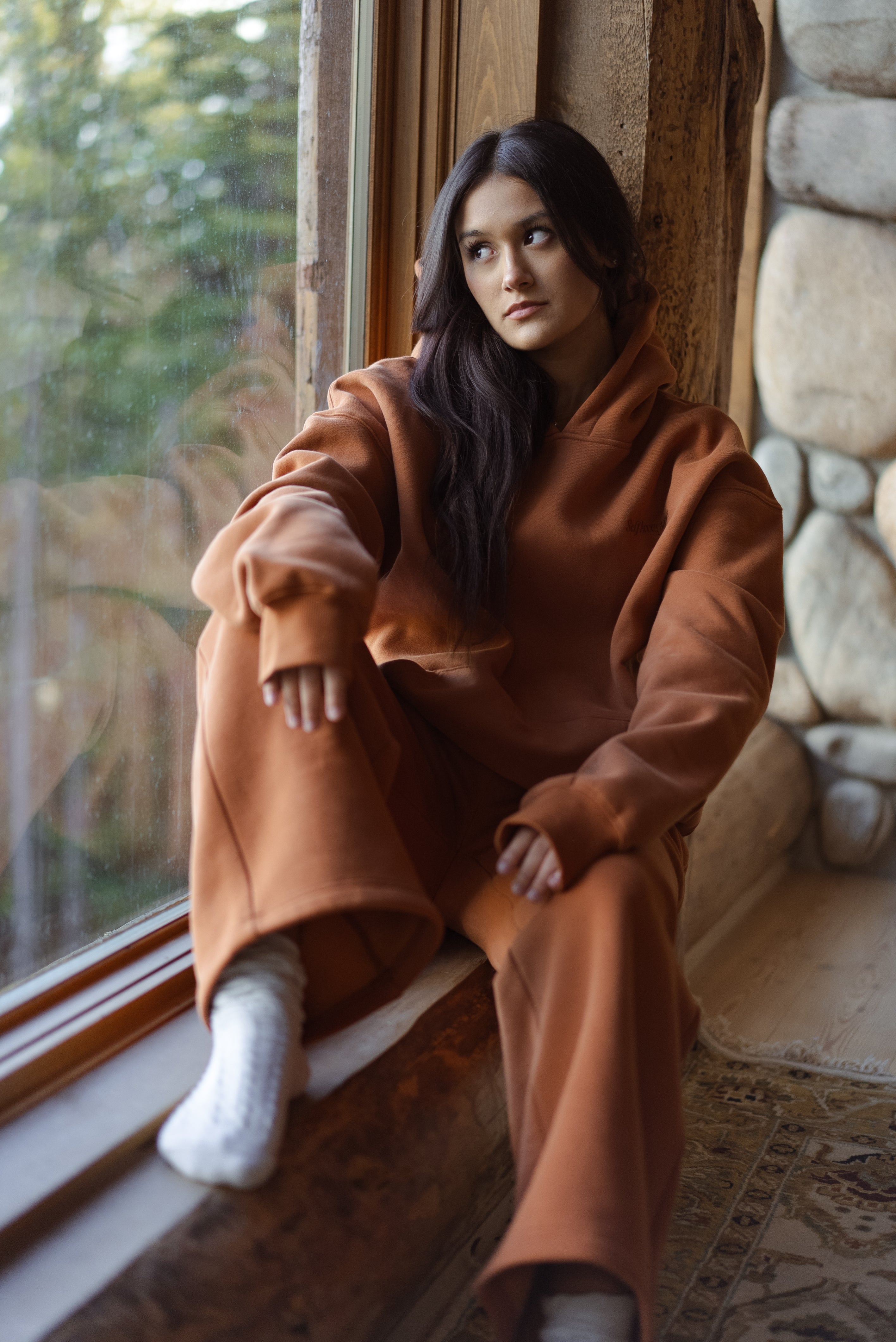 Woman wearing a Pumpkin Spice Self Acceptance™ hoodie and straight-leg sweatpants while sitting beside a cabin window.